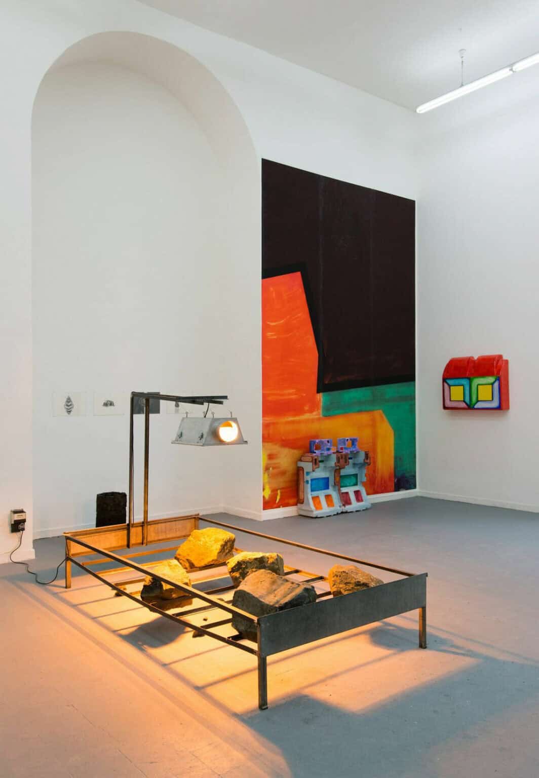 Foto: Ein Raum mit weißen Wänden, grauem Boden, einem großen Fenster und einer wandhohen, bogenförmigen Einbuchtung. An den Wänden befinden sich mehrere Objekte, einige von ihnen sind plastisch, andere flach. Sie hängen auf unterschiedlichen Höhen. Im Raum befindet sich eine Skulpturen. Ein Licht ist Teil der Skulptur. Es ist eingeschaltet und taucht Teile des Raums in warmes Licht und lässt das Objekt einen Schatten werfen.