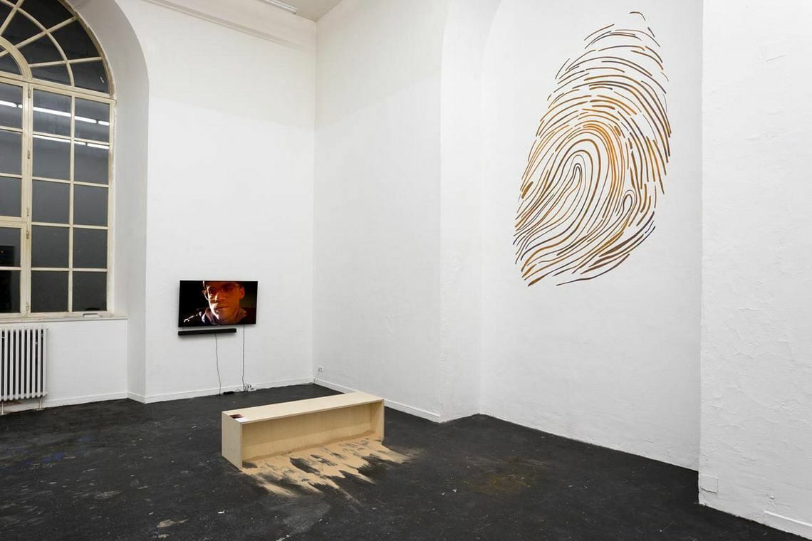 Foto: Ein Raum mit weißen Wände, schwarzem Boden und einem großen Fenster. In einer bogenförmigen Einbuchtung an der Wand befindet sich eine Wandbemalung. An einer der Wände hängt ein Bildschirm. Im Raum steht ein rechteckiges Objekt auf dem Boden, um das herum Sand liegt.