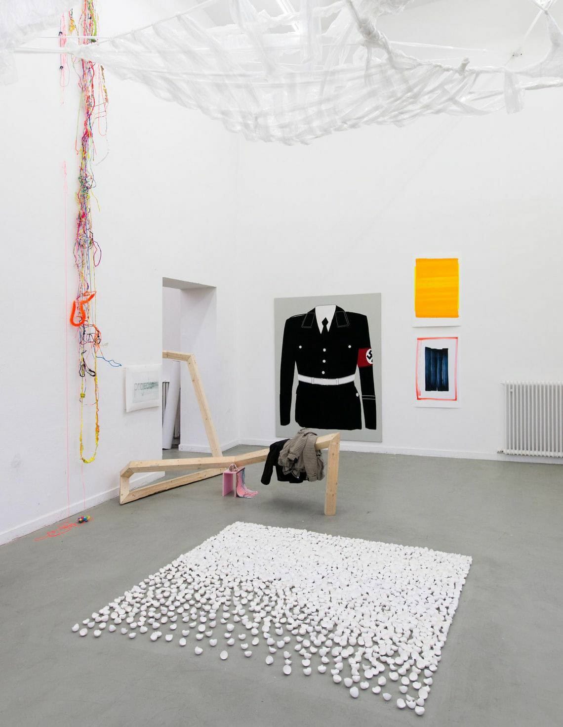 Foto: Ein Raum mit weißen Wänden und grauem Boden. n den Wänden befinden sich mehrere Objekte, einige von ihnen sind plastisch, andere flach. Sie hängen auf unterschiedlicher Höhe. Im Raum befinden sich mehrere Skulpturen auf unterschiedlichen Materialien direkt auf dem Boden.