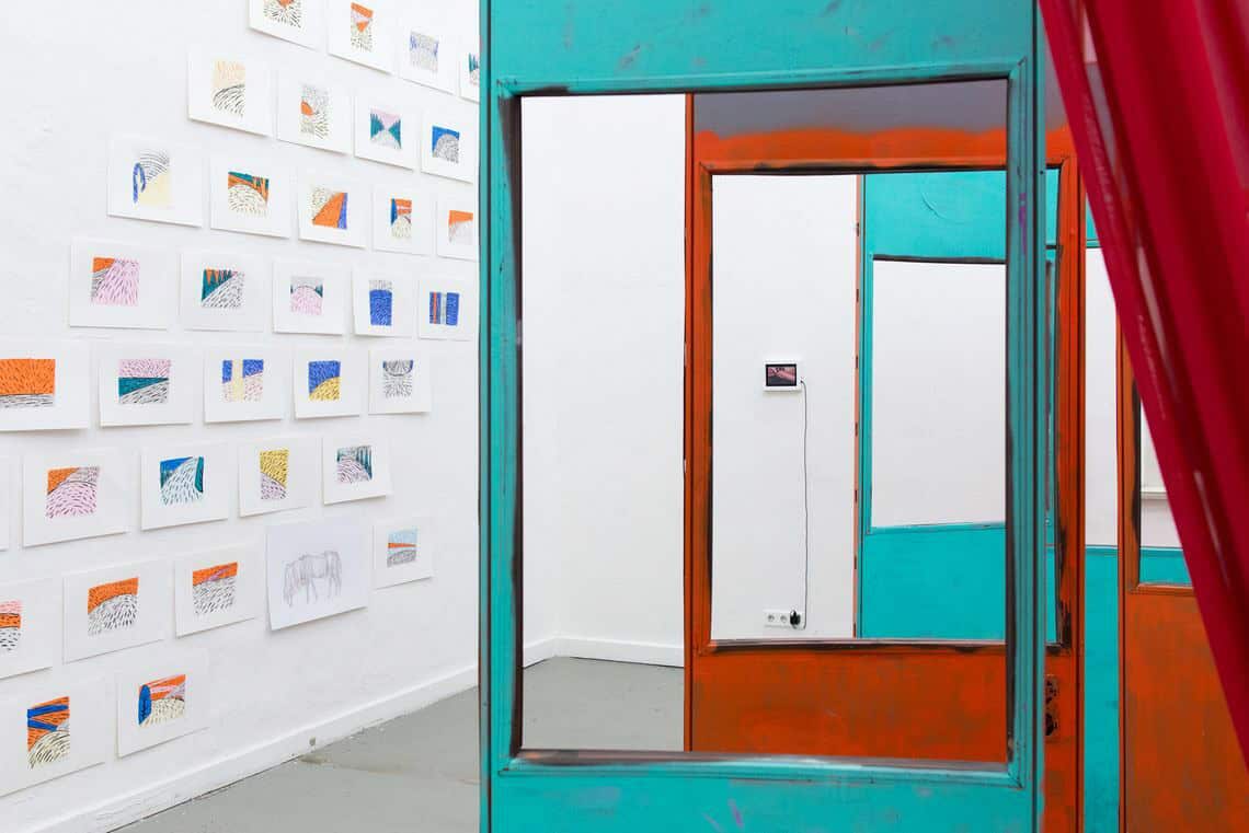 Foto: Ein Raum mit weißen Wänden, grauem Boden und einem großen Fenster. An den Wänden hängen mehrere flache Objekte neben- und untereinander. Auch ein kleiner Bildschirm hängt an der Wand. Im Raum befindet sich eine Skulptur.