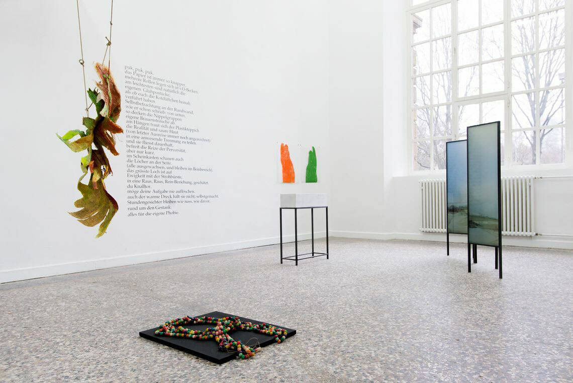 Foto: Ein Raum mit weißen Wänden, mehrfarbigem Steinboden und einem großen Fenster. An der Wand befindet sich ein Text und flache Objekte. Im Raum Stegen Objekte. Auch von der Decke hängt ein Objekte. Die Dinge sind auf unterschiedlichen Materialien gefertigt.