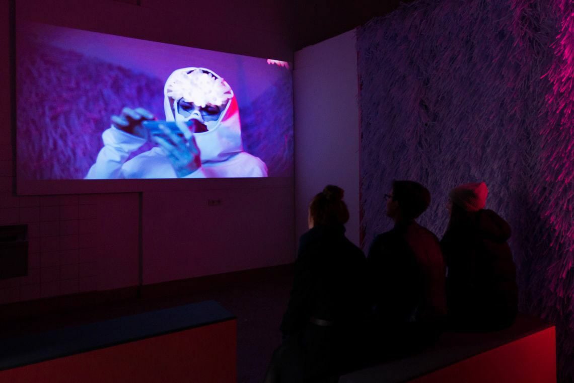 Foto: Ein abgedunkelter Raum der in rötlich-lilanes Licht getaucht ist. Im Raum befindet sich eine Projektion und einige Menschen.