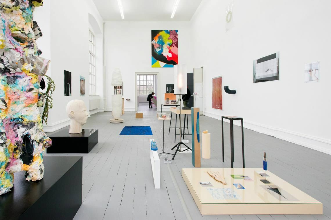 Foto: Ein Raum mit weißen Wänden, grauem Boden und großen Fenstern. An den Wänden befinden sich mehrere Objekte, einige von ihnen sind plastisch, andere flach. Sie hängen auf unterschiedlicher Höhe. Auf dem Boden liegen und stehen viele Objekte. Manche von ihnen sind zu mehreren auf flächigen Sockeln arrangiert, andere direkt auf dem Boden angebracht. Die Elemente stehen zum Teil sehr eng beieinander. Hinten im Raum befindet sich eine Person.