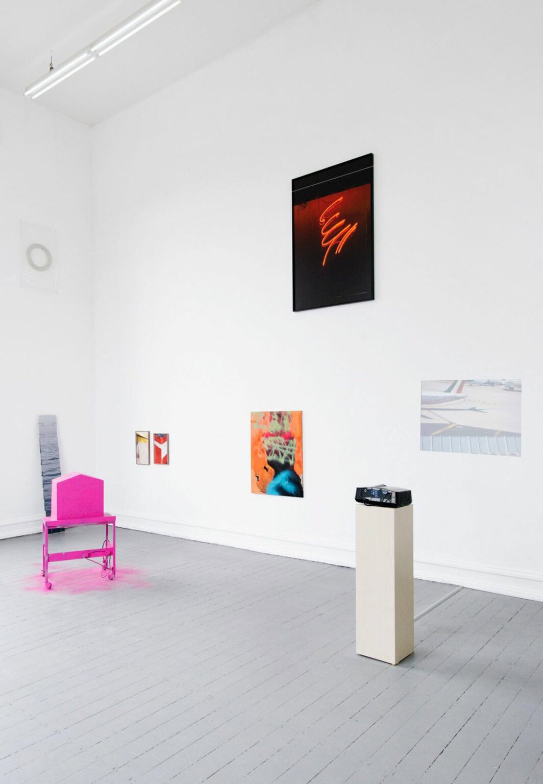 Foto: Ein Raum mit weißen Wänden und grauem Dielenboden. An den Wänden hängen mehrere flache Objekte neben- und untereinander. Manche von Ihnen sind gerahmt. Im Raum befinden sich Skulpturen, die auf dem Boden stehen oder an der Wand lehnen. Vor einer der Wände steht ein Sockel, auf dem ein Beamer steht. Ein wirft eine Projektion an die Wand.