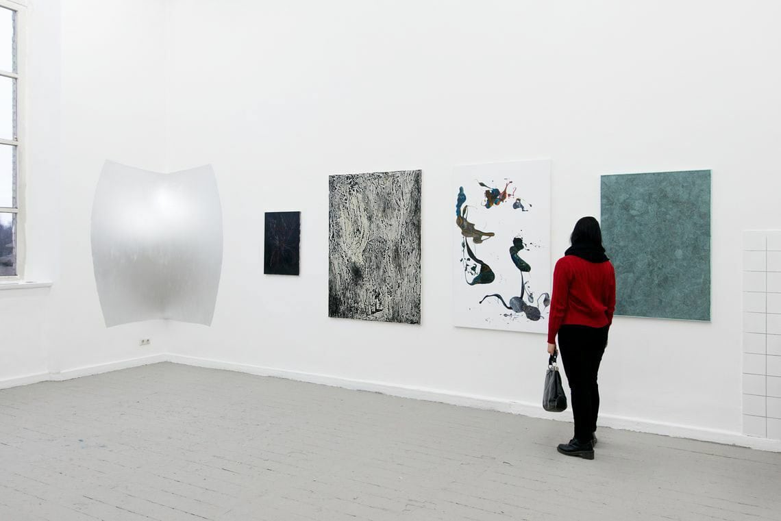 Foto: Ein Raum mit weißen Wänden, grauem Boden und einem großen Fenster. An den Wänden hängen mehrere Leinwände und Objekte auf ähnlicher Höhe nebeneinander. Im Raum befindet sich ein Mensch.