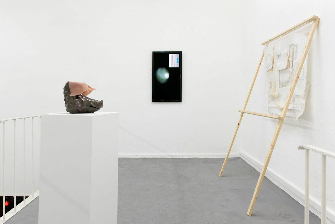 Foto: Ein Raum mit weißen Wänden und grauem Boden. An der Wand hängt ein Flaches Objekt. Im Raum stehen Objekte auf dem Boden oder auf Sockeln. Sie sind auf unterschiedlichen Materialien gefertigt.