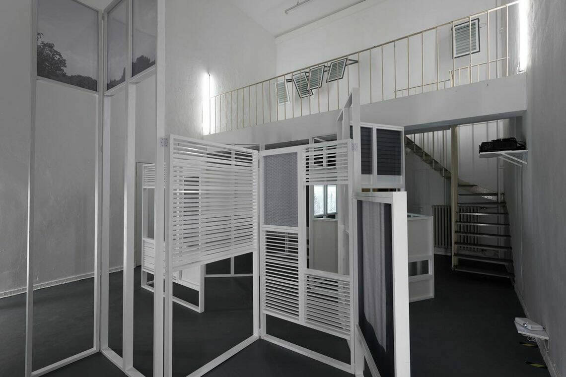 Foto: Ein Raum mit weißen Wänden, dunkelgrauem Boden, und einer Empore, die durch eine Wendeltreppe erreicht werden kann. Im Raum befindet sich eine verschachtelte Konstruktion, in denen verschiedene Materialien, unter anderem Lamellen-Vorhang artige Strukturen verbaut sind. An der Wand befindet sich ebenfalls Objekte.