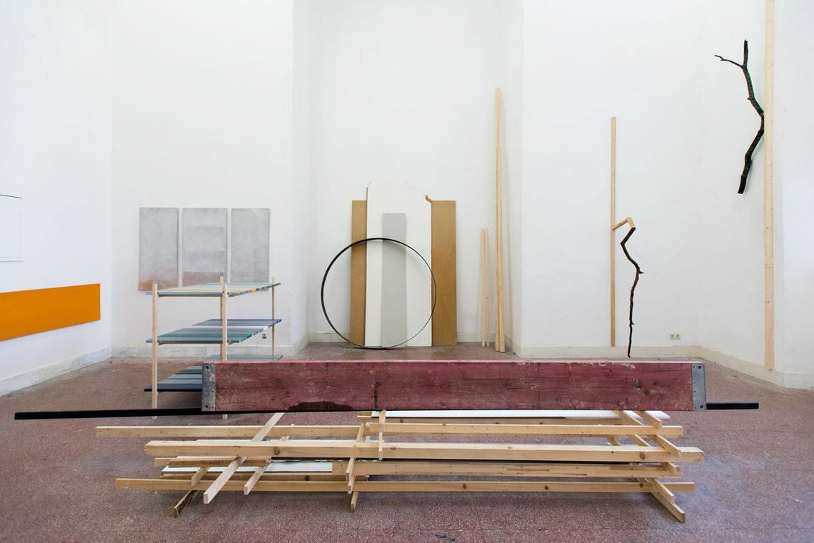 Foto: Ein Raum mit weißen Wänden, rotem Steinboden und einer wandhohen Einbuchtung. An der Wand des Raumes, in der Einbuchtung und im Raum hängen, stehen, lehnen und liegen Objekte auf unterschiedlichen Materialien.