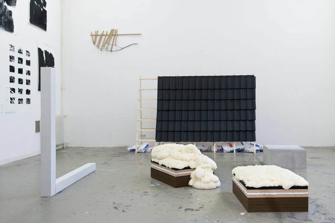 Foto: Ein Raum mit weißen Wänden und grauem Boden. Der Boden ist mit Farbsprencklern übersäht. An den Wänden hängen mehre Flache Objekte in unterschiedlichen Materialien auf unterschiedlicher Höhe. Im Raum stehen mehrere Skulpturen, zum teil nah beieinander.