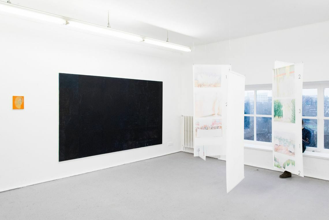 Foto: Ein Raum mit weißen Wänden, grauem Boden und einem kleinen Fenster. An der Wand des Raumes hängen zwei Leinwände in Klein- und Großformat. Von der Decke hängen drei bemalte Bögen.