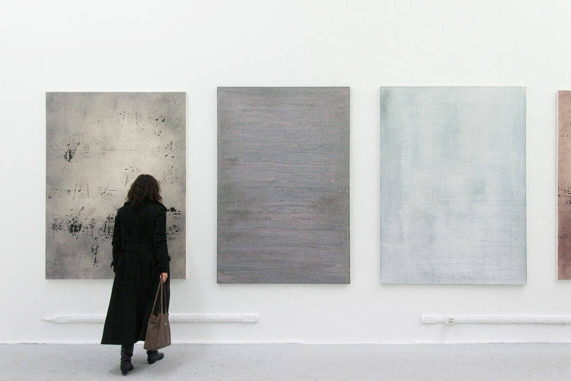 Foto: Ein Raum mit weißen Wänden und grauem Boden. An der Wand mehrere große Leinwände im ähnlichen Format, die auf gleicher Höhe hängen. Im Raum befindet sich eine Person.