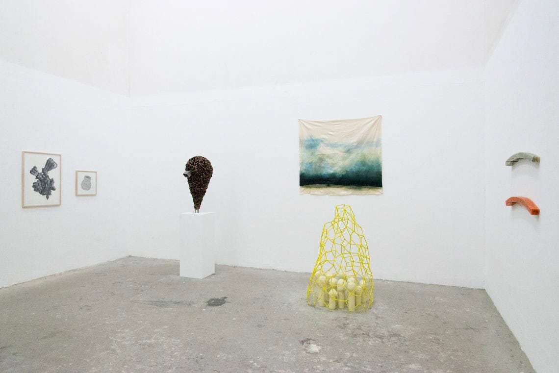 Foto: Ein Raum mit weißen Wänden und grauem Boden. An den Wänden hängen flache, zum Teil gerahmte Objekte. Im Raum stehen zwei Skulpturen, eine von ihnen auf einem Sockel, die andere auf dem Boden.