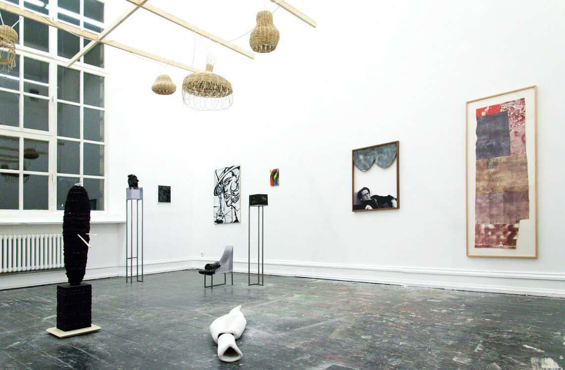 Foto: Ein Raum mit weißen Wänden, dunklem, mit bunten Farbflecken bedecktem Boden und einem großen Fenster. An den Wänden befinden sich mehrere, zum Teil gerahmte, flache Objekte. Im Raum stehen und liegen mehrere Skulpturen. Von der Decke hängt eine Konstruktion. Die Objekte sind aus unterschiedlichen Materialien gefertigt.