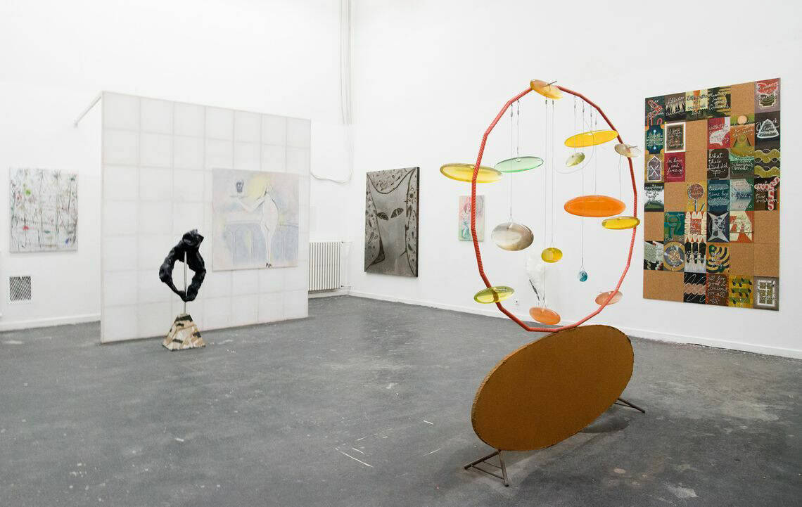 Foto: Ein Raum mit weißen Wänden und dunklem Boden. An den Wänden befinden sich mehrere flache Objekte. Im Raum stehen mehrere Skulpturen. Unweit einer der Wände ist eine weiße Wand eingezogen, die an der dahinterliegende Wand befestigt ist. Auf der Fläche ist eine Leinwand angebracht.