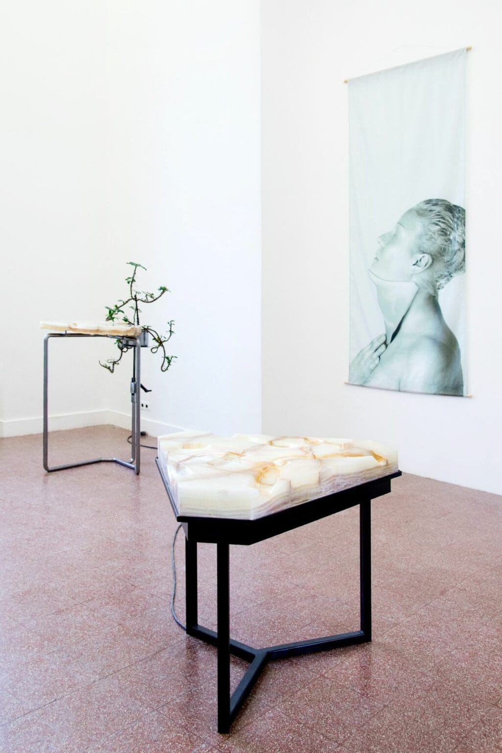 Foto: Ein Raum mit weißen Wänden, rotem Steinboden und einer wandhohen Einbuchtung. Im Raum befinden sich zwei Skulpturen. An der Wand, innerhalb der Einbuchtung, ein flaches Objekt im Hochformat.