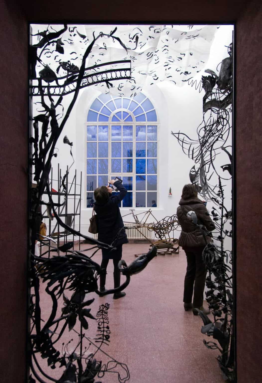 Foto: Ein Raum mit weißen Wänden, rotem Steinboden und einem großen Fenster. Im gesamten Raum befinden sich Skulpturen. Einige von ihnen ragen in den Türrahmen herein. Im Raum befinden sich außerdem zwei Menschen.