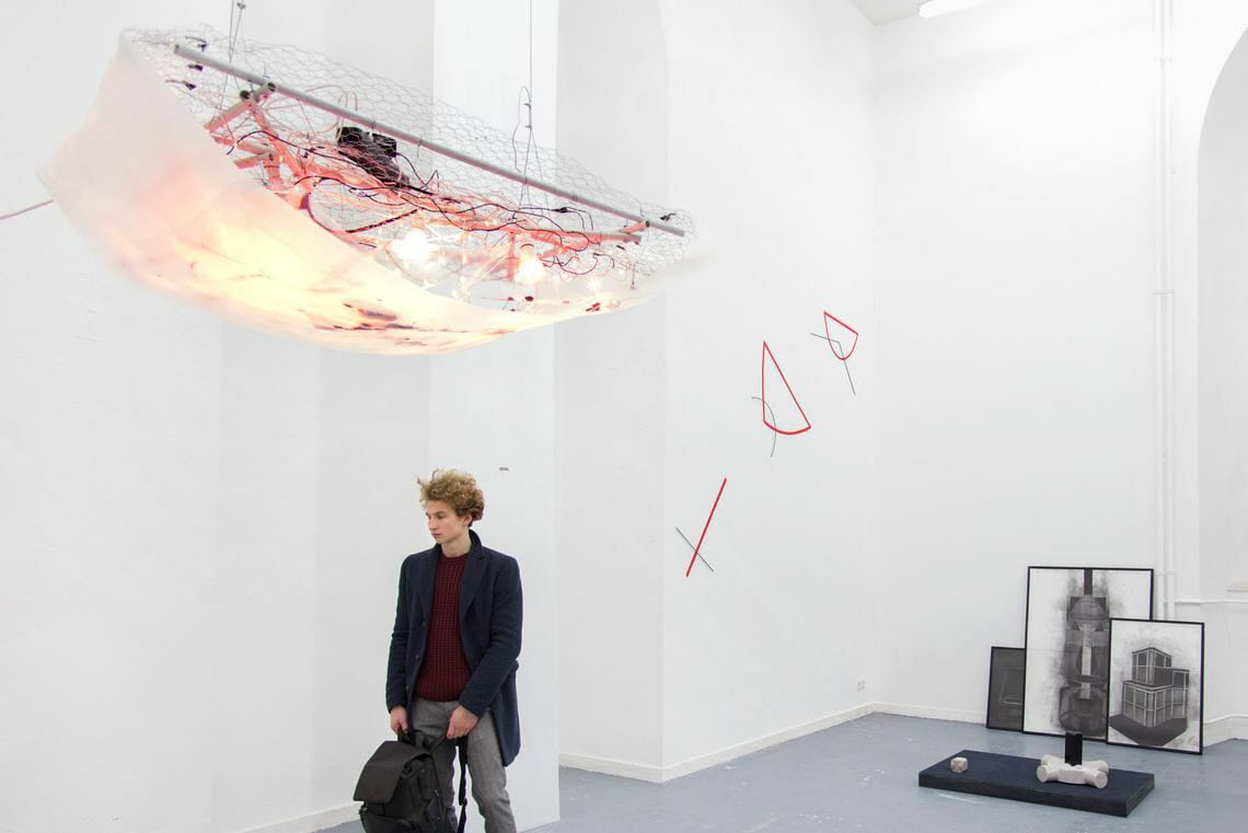 Foto: Ein Raum mit weißen Wänden, grauem Boden und zwei wandhohen, bogenförmigen Einbuchtungen. Von der Decke hängt ein großes Objekt, in das leuchtende Lichter integriert sind. Unter dem Objekt steht ein Mensch. Weit oben an der Wand befindet sich eine Gruppe flacher Objekte. Im Raum auf dem Boden liegen und an der Wand lehnend befinden sich Objekte, die aus unterschiedlichen Materialien bestehen.