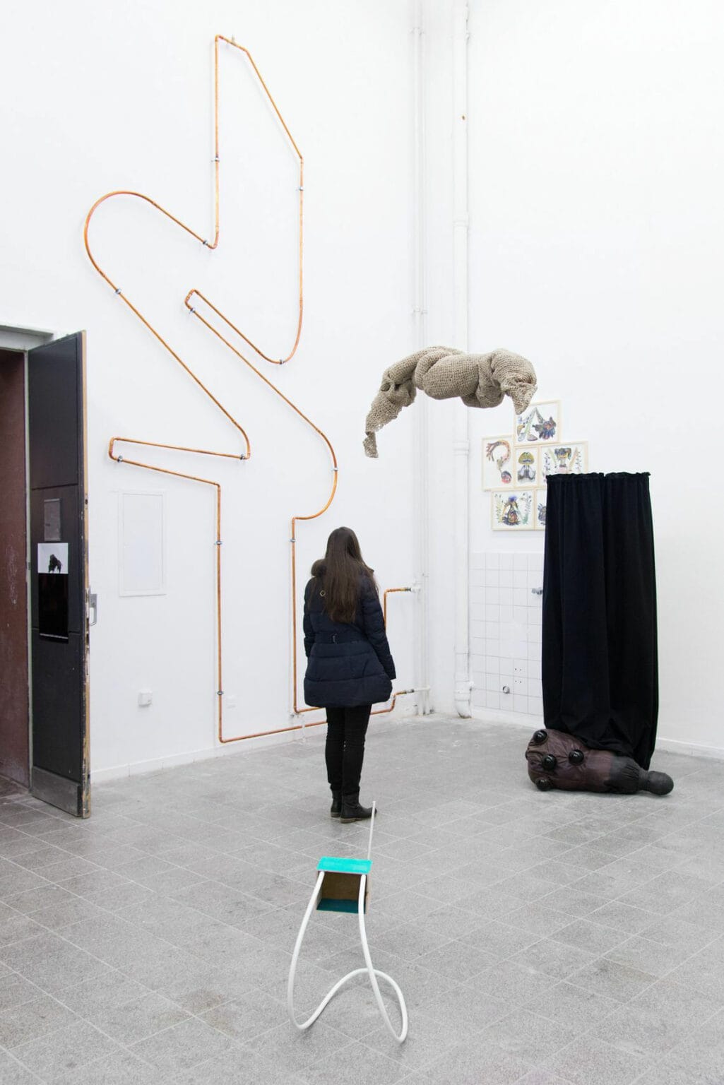 Foto: Ein Raum mit weißen Wänden, grauem, gefließten Steinboden und einer Tür. Im Raum befinden sich Skulpturen, die zum teil auf dem Boden liegen, zum Teil frei im Raum stehen oder von der Decke hängen. Sie sind aus unterschiedlichen Materialien gefertigt. An der Wand ist ein großes Objetkt und mehrere kleine gerahmte Objekte montiert. Im Raum befindet sich ein Mensch.