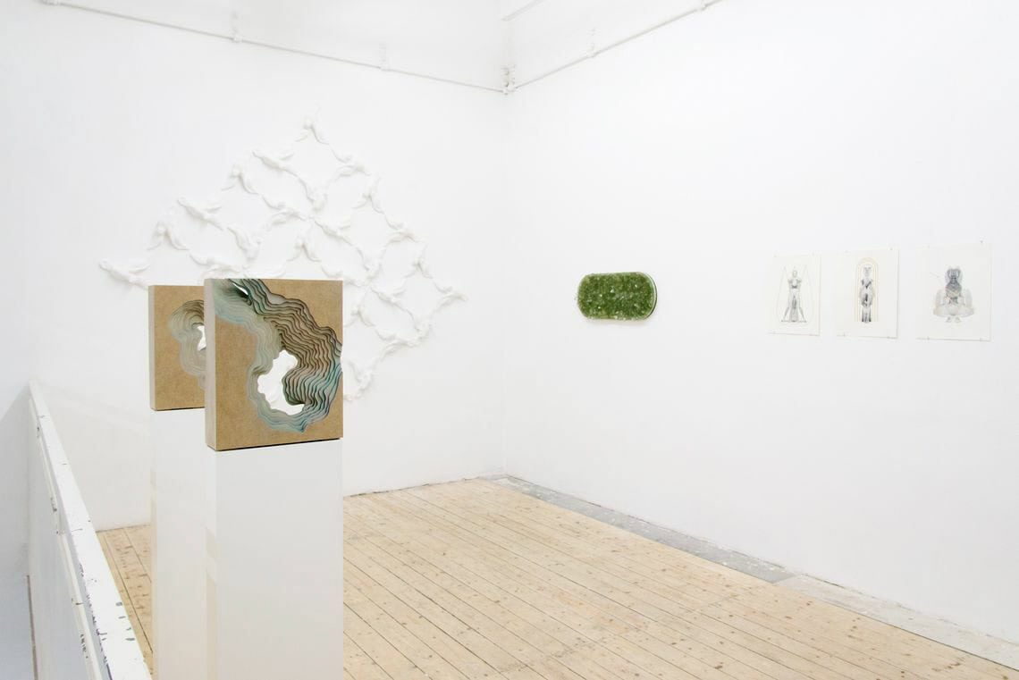 Foto: Ein Raum mit weißen Wänden und Dielenboden. An den Wänden hängen mehrere flache Objekte und Papiere. Im Raum stehen Objekte auf Sockeln.