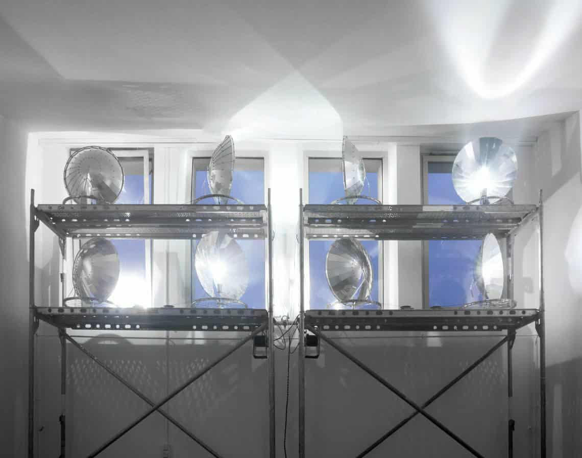Foto: Ein Raum mit weißen Wänden und mehreren kleinen Fenstern, die sich oben an der Wand befinden. Vor der Wand ist eine Metallkonstruktion aufgebaut. Auf den Boden der Metallkonstruktion stehen silberne, reflektierende Schüsseln, die teile des Raums erleuchten.