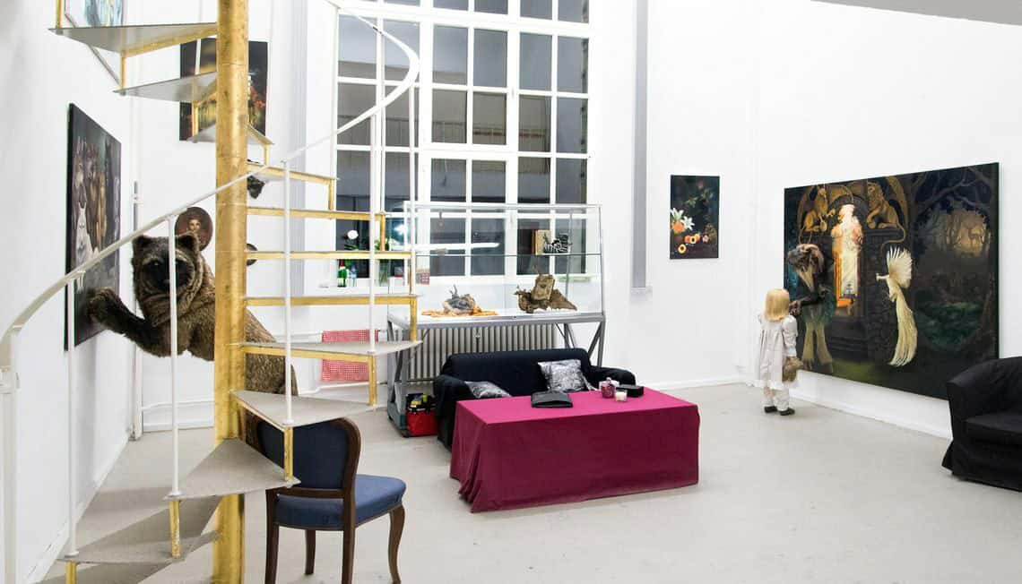 Foto: Ein Raum mit weißen Wänden, grauem Boden und einer grau-goldenen Wendeltreppe. Im Raum befinden sich mehrere Objekte, darunter auch Möbel und gefüllte Vitrinen. An den Wänden befinden sich auf unterschiedlicher Höhe hängend Malereien.