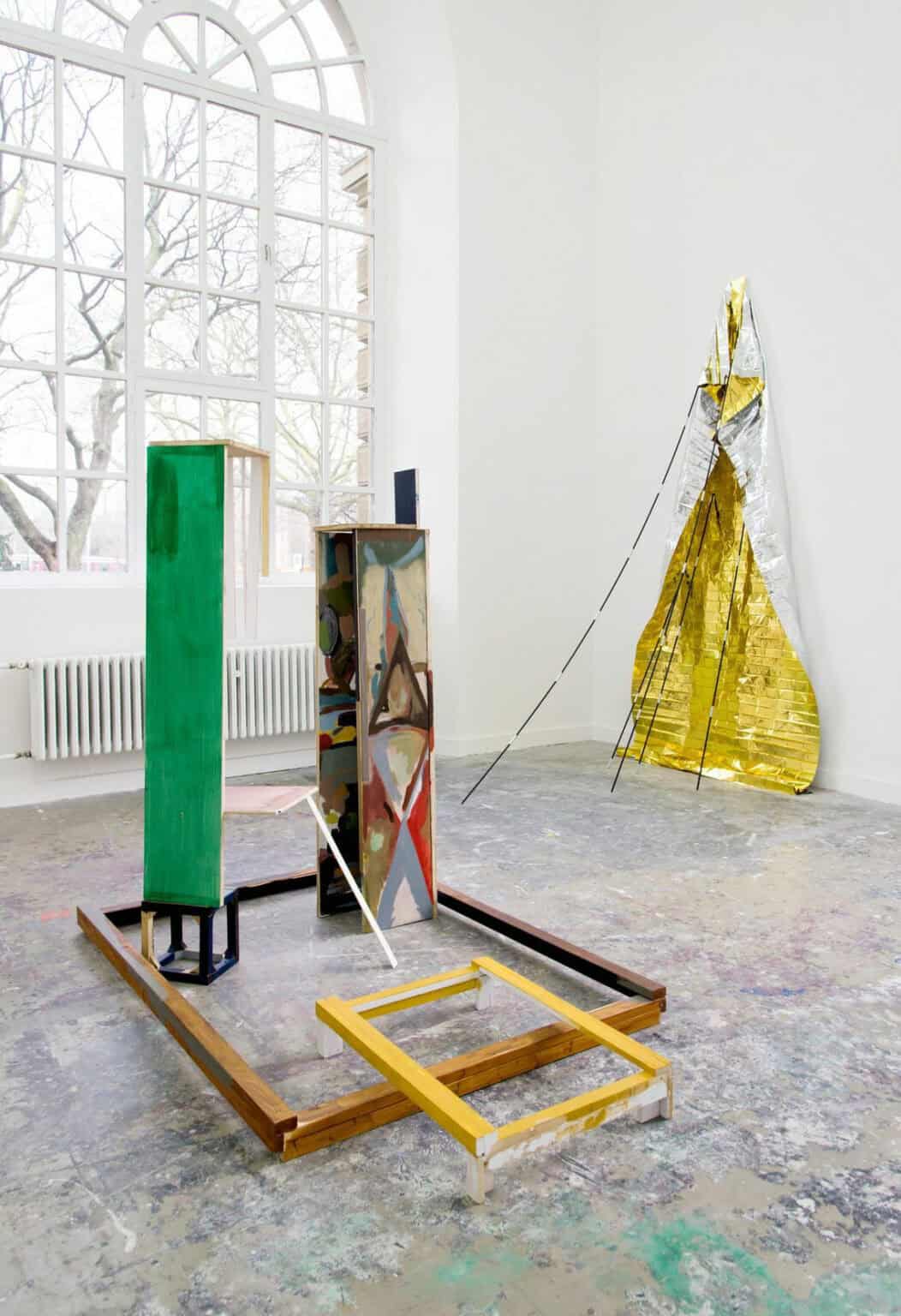 Foto: Ein Raum mit weißen Wänden, gräulich-buntem Boden und einem großen Fenster. An der Wand und im Raum befinden sich große Skulpturen aus unterschiedlichen Materialen. An der Wand nahe des Fensters hängt ein kleinformatiges, flaches Objekt.