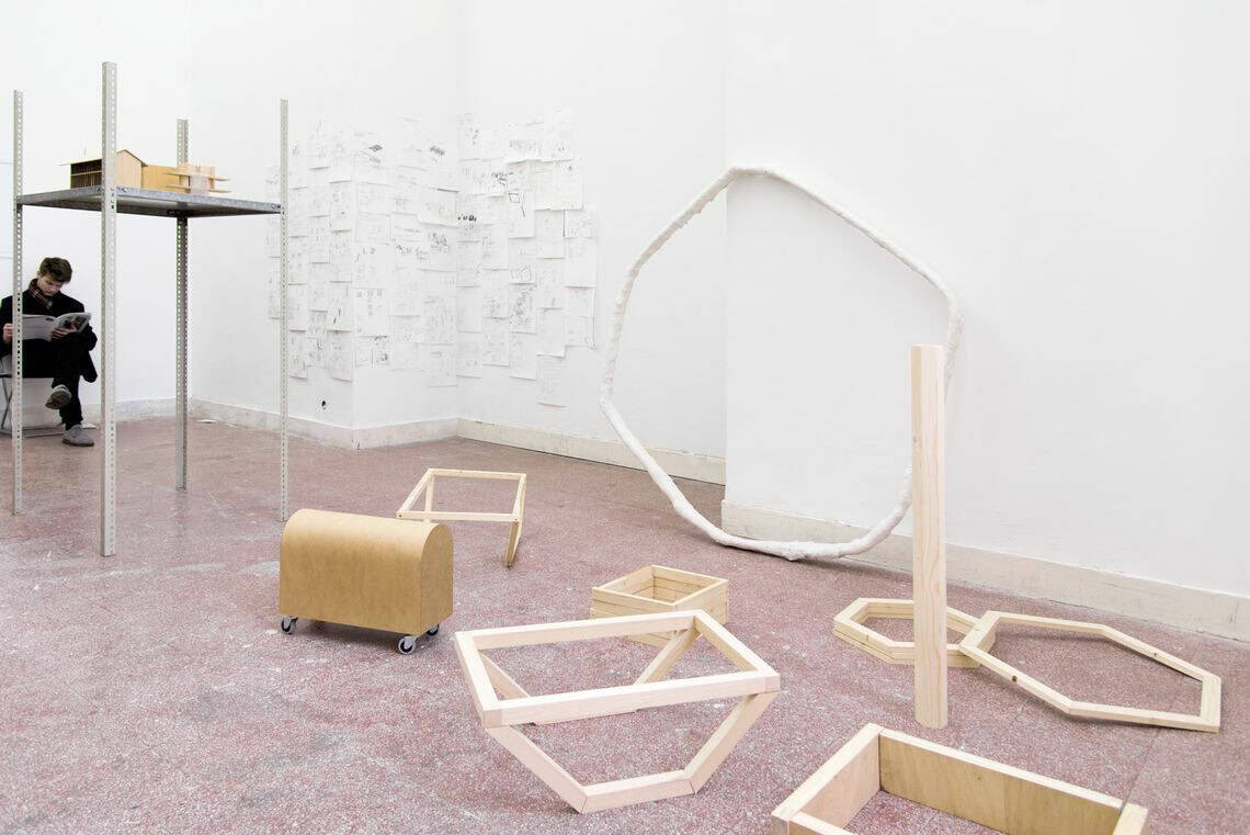 Foto: Ein Raum mit weißen Wänden, rotem Steinboden und einer Wandhohen, bogenförmigen Einbuchtung. Im Raum befinden sich mehrere Objekte aus unterschiedlichen Materialien, die nah beieinander liegen. An der Wand befindet sich ein Arrangement auf bemalten Papieren in A4 im Hoch- und Querformat.