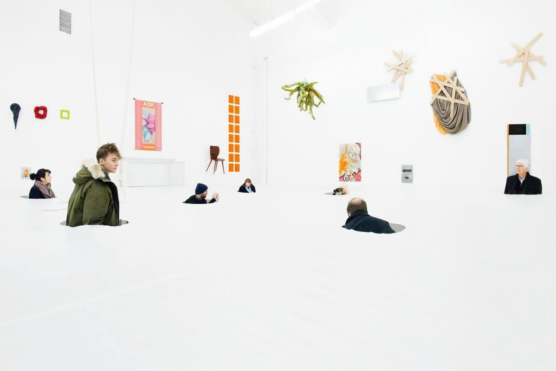 Foto: Ein Raum mit weißen Wänden und weißen Boden. In dem Boden sind Löcher, auf dem die Köpfe und Oberkörper von Menschen in den Raum herein ragen. An den Wänden hängen viele teils flache, teils plastische Objekte.