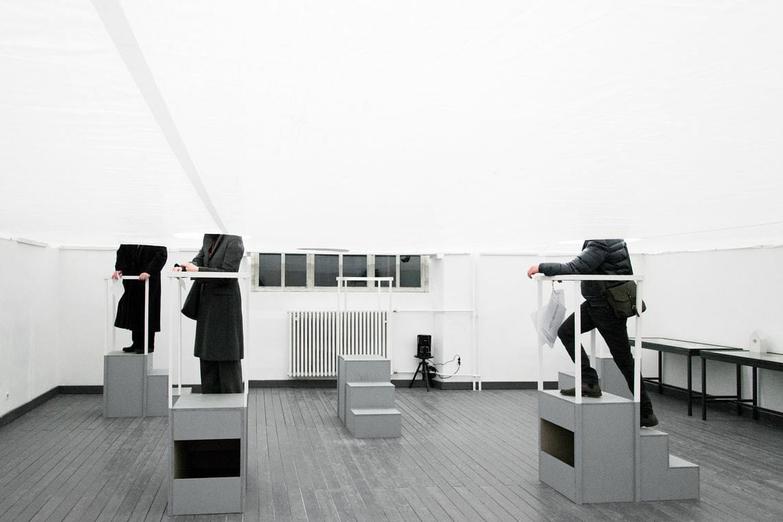 Foto: Ein Raum mit weißen Wänden, grauem Dielenboden und einem Fenster. Mittig im Raum ist eine weiße Plane gespannt. Im Raum stehen treppenartige Podeste und Tische. Menschen stehen auf den Podesten. Ihr Kopf verschwindet in Löchern, die sich überhalb der Podeste in der Plane befinden. Somit sind nur ihre Unterkörper zu sehen.