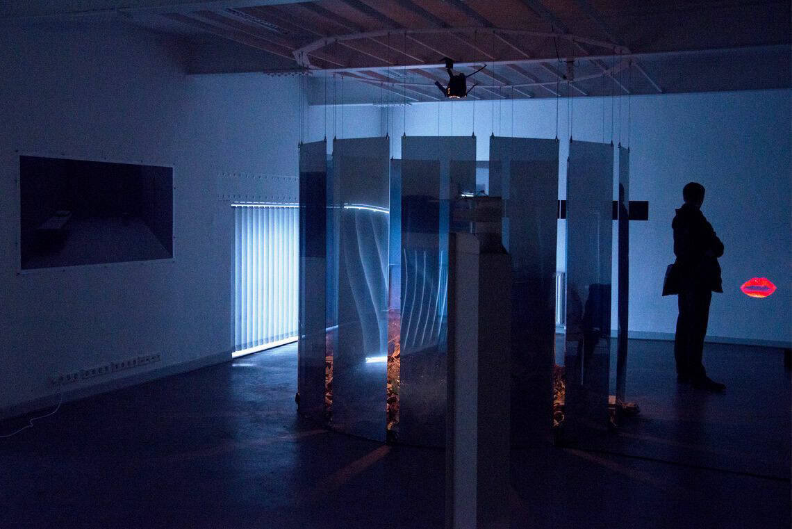 Foto: Ein abgedunkelter Raum. Die Fenster sind mit Lamallen-Vorhängen bedeckt, durch die ein bläuliches Licht dringt. An den Wänden des Raumes hängt ein flaches Objekt und an eine andere Wand wird eine kleine Projektion geworfen. Im Raum steht eine Skulptur. Von der Decke des Raumes hängen in kreisförmiger Anordnung halbtransparente Platten. Innerhalb der Platten liegt etwas auf dem Boden, das von einem warmen Licht angestrahlt wird. Im Raum befindet sich ein Mensch.