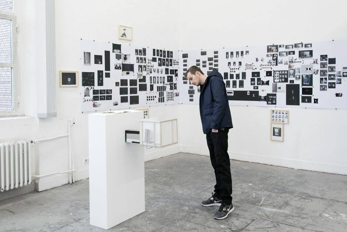 Foto: Ein Raum mit weißen Wänden, grauem Boden und einem großen Fenster. An den Wänden den befinden sich mehrere flache Objekte, manche von Ihnen sind gerahmt. Im Raum steht eine Skulptur und ein Mensch.