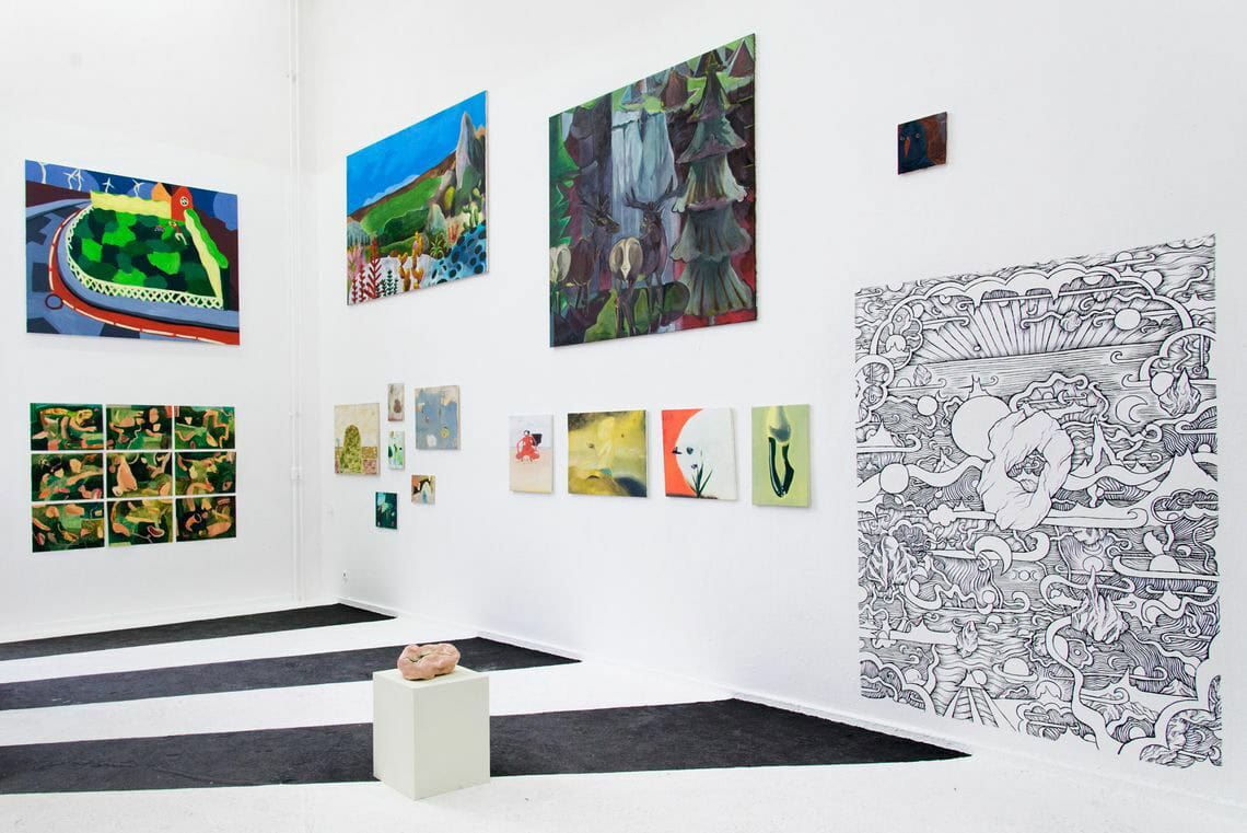 Foto: Ein Raum mit weißen Wänden und schwarz weißen, gestreiften Boden. An den Wänden hängen neben- und untereinander mehrere flache Objekte in Klein-, Mittel- und Großformaten. Auf dem Boden steht ein niedriger Sockel auf dem sich eine Skulptur befindet.