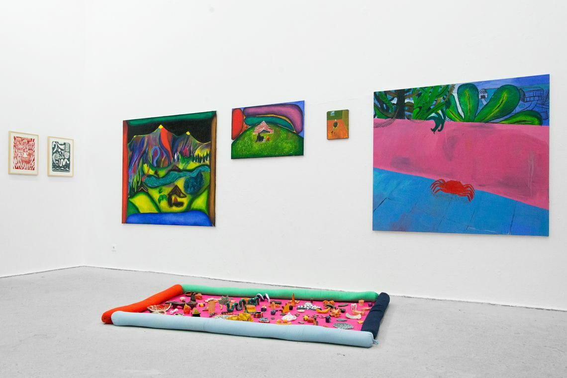 Foto: Ein Raum mit weißen Wänden und grauem Boden. An der Wand befinden sich mehrere Leinwände im Klein- und Mittelfromat und zwei kleine, gerahmte Objekte. Auf dem Boden liegt ein Arrangement aus Objekten.