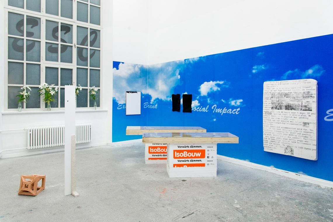 Foto: Ein Raum mit weißen Wänden, grauem Boden und einem großen Fenster. Ein Teil der Wand ist mit einem brauen Material Bedeckt, auf den Wolken und ein Schriftzug gedruckt sind. Auf der blauen Fläche sind mehrere flache Ibkekte angebracht. Auf der Fensterbank stehen mehrere Blumen in Wasen. Das dahinterliegende Fenster ist mit milchigen Folien und einem Schriftzug beklebt. Im Raum stehen mehrere Skulpturen.