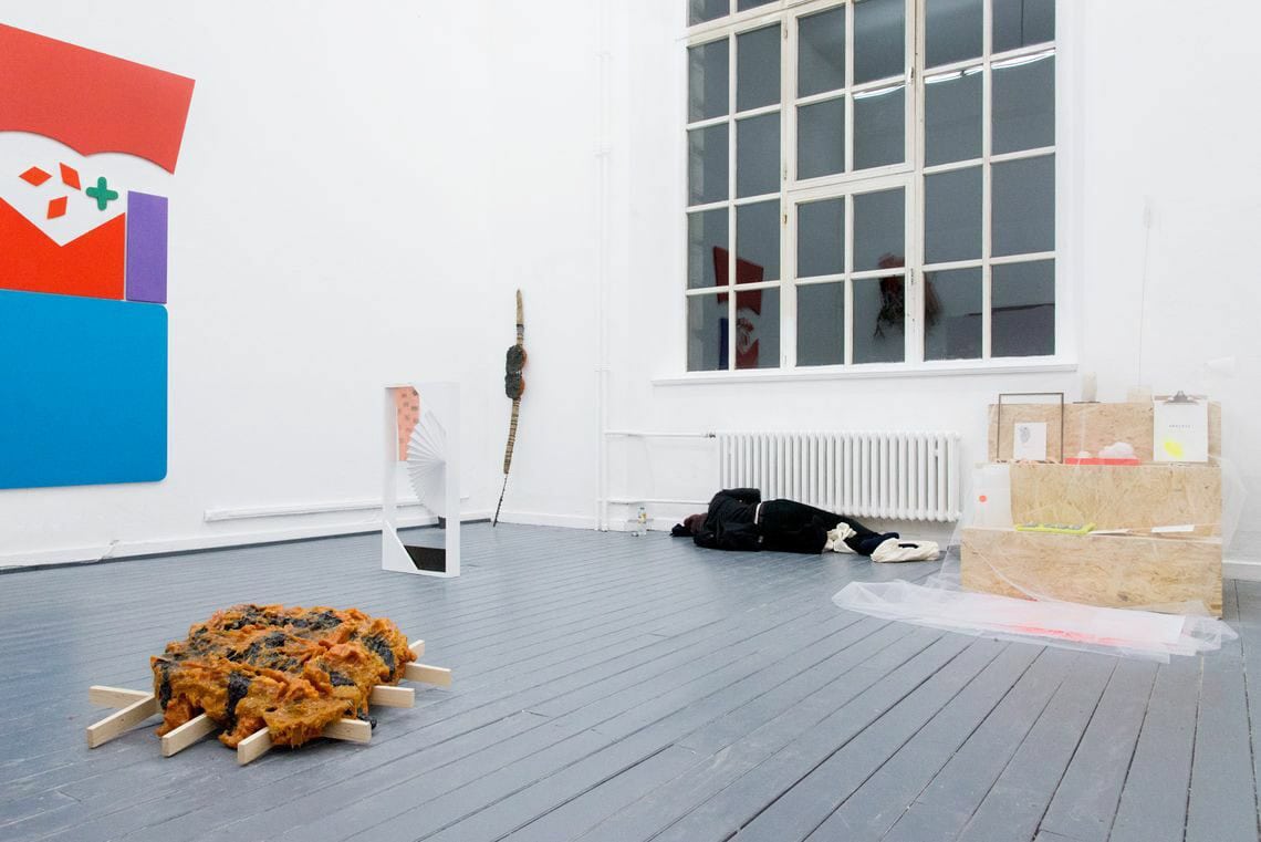 Foto: Ein Raum mit weißen Wänden, grauem Dielenboden und einem großen Fenster. An der Wand befindet sich ein großes, flaches Objekt. Auf dem Boden des Raumes stehen und lehnen Objekte auf unterschiedlichen Materialien.