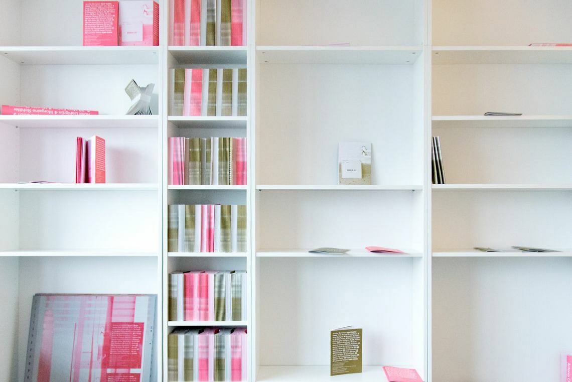 Foto: Mehrere, eng aneinander stehende weiße Regale in unterschiedlichen Breiten. Die Regalbretter sind zum Teil auf unterschiedlicher Höhe angebracht. Auf den Regalbrettern stehen und liegen Hefte in den Farben rosa und grün.