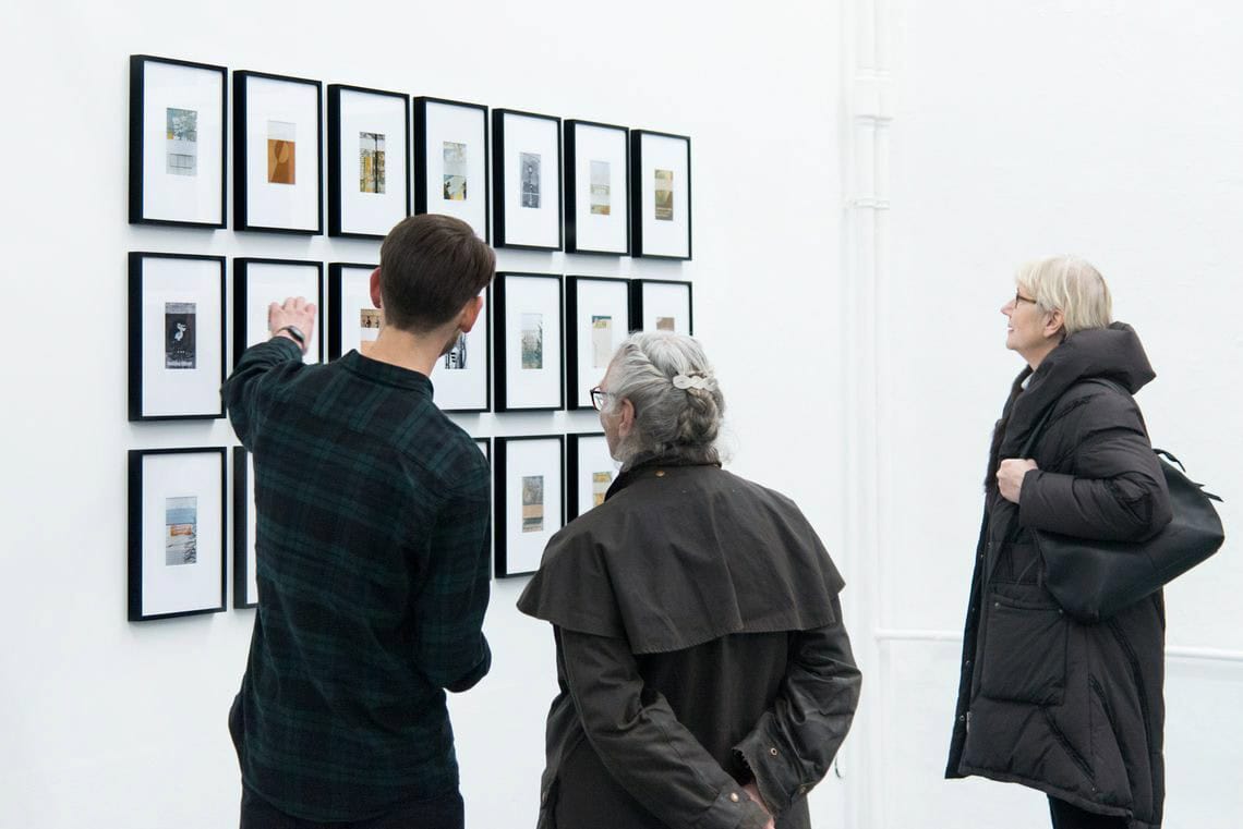 Foto: Ein Raum mit weißen Wänden. An der Wand hängen mehrere gerahmte Objekte in drei Reihen rasterartig neben- und untereinander. Um Raum befinden sich Menschen.