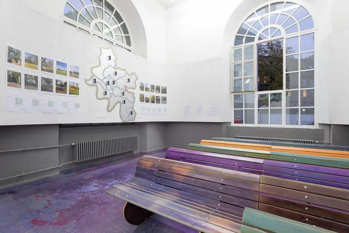 Foto: Ein Raum mit weißen Wänden und zwei großen Bogenfenstern. Der Boden ist in einem fleckigen lila-blau Ton bemalt. Der untere Teil der Wand ist mit einem grauen streifen bemalt. Teile des Fensters sind vertäfelt. Auf der Vertäfelung und auf der Wand befinden sich flache Objekte und bedruckte Zettel. Im Raum steht eine flächige Konstruktion.