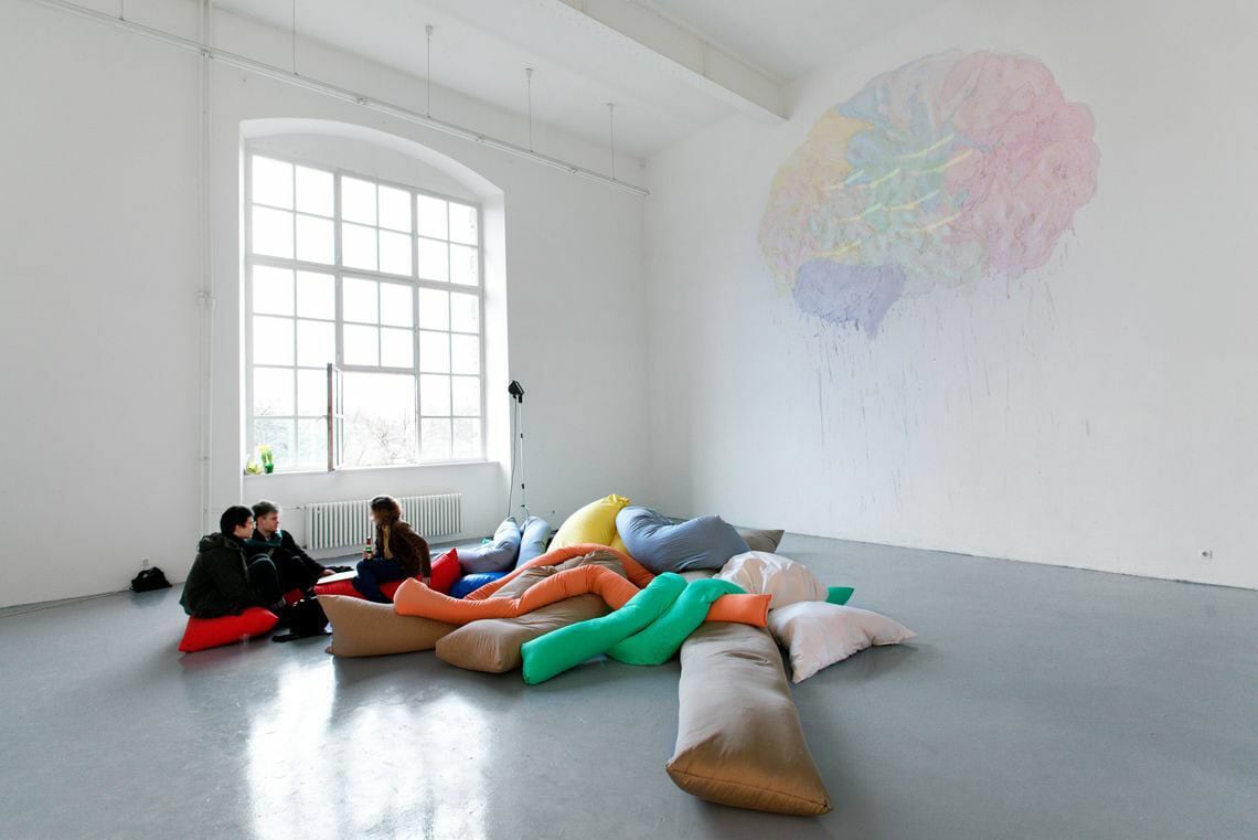 Foto: Ein Raum mit weißen Wänden, grauem Boden und einem großen Fenster. Hoch oben ist die Wand mit unterschiedlichen, blassen Farben bemalt. Im Raum befindet sich ein Haufen an Objekten, auf dem Menschen sitzen.