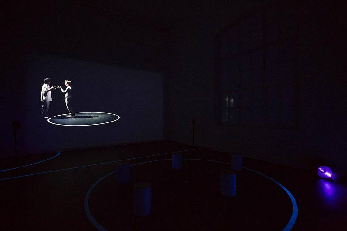 Foto: Ein dunkler Raum, in dem ein lila leuchtendes Licht scheint. An der Wand eine Projektion.