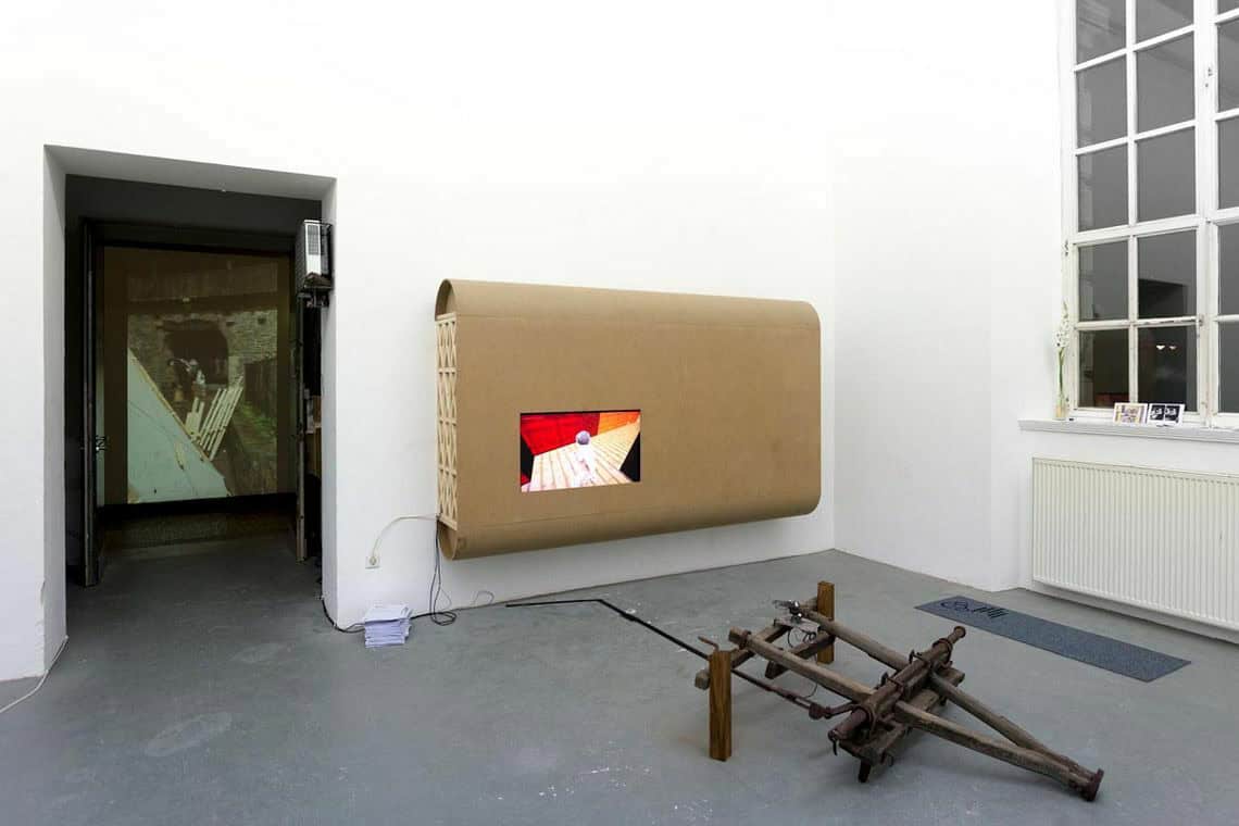 Foto: Ein Raum mit weißen Wänden, grauem Boden, einem Fenster und einer Tür. An der Wand befindet sich eine Konstruktion aus unterschiedlichen Materialien, auf dem Boden liegen Objekte. Durch die Türöffnung hindurch wird eine Projektion auf eine Wand geworfen.