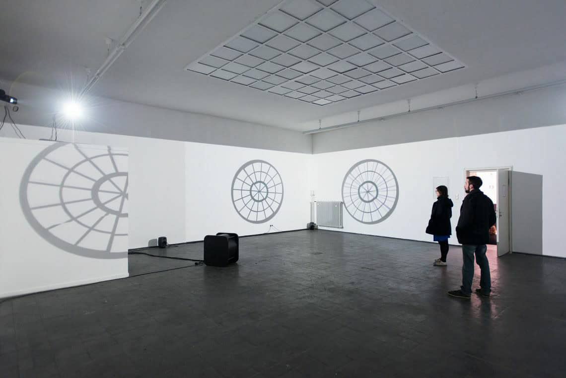 Foto: Ein Raum mit weißen Wänden. Auf die Wände werden mehrere Projektionen in unterschiedlichen Formaten geworfen. Im Raum befinden sich Menschen.