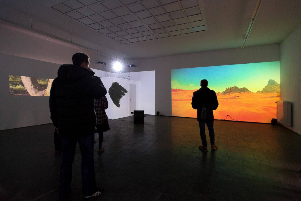 Foto: Ein Raum mit weißen Wänden. Auf die Wände werden mehrere Projektionen in unterschiedlichen Formaten geworfen. Im Raum befinden sich Menschen.