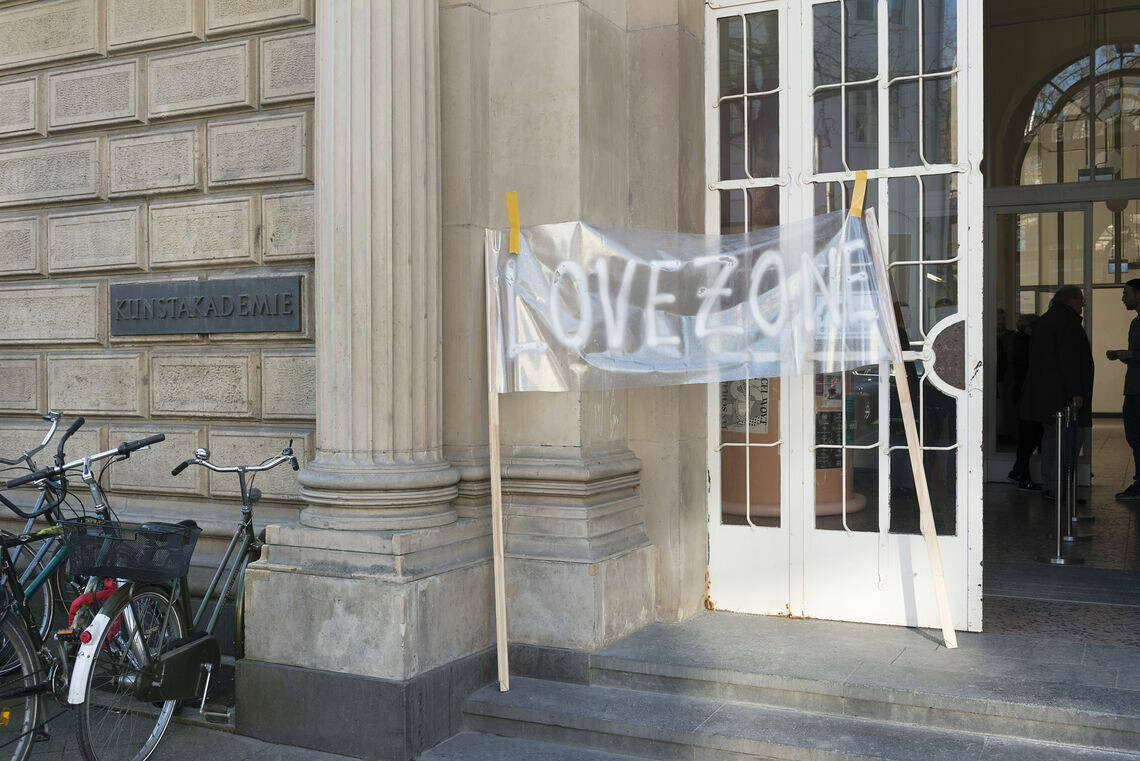 Foto: Dir Tür eines Gebäudes mit Steinfassade und Säulenelementen. Ein Flügel der Tür ist geöffnet. Auf der Fassade ist ein Schild mit dem Wort "KUNSTAKADEMIE" befestigt. Im Eingangsbereich lehnen zwei Holzleisten, zwischen denen ein transparentes Banner befestigt ist, auf dem "LOVE ZONE" steht.