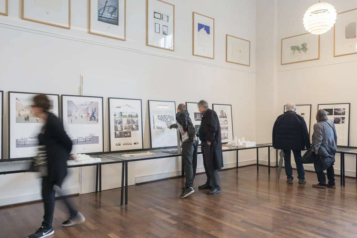 Foto: Ein Raum mit weißen Wänden und braunem, poliertem Dielenboden. Oben an der Wand befindet sich eine Reihe gerahmter Objekte. Wandnah stehen mehrere Tische des gleichen Modells, vor denen sich Menschen bewegen. Auf den Tischen liegen modellartige Objekte. Auf ihnen stehen außerdem gefüllte Bilderrahmen, die an der Wand lehnen.