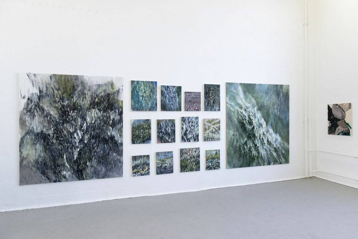 Foto: Ein Raum mit weißen Wänden und grauem Boden. An den Wänden hängt eine Gruppe kleinformatiger Leinwände in einem Raster über- und nebeneinander .Sie sind von zwei großformatigen Leinwänden eingeklammert. An einer weiteren Wand hängt ein weiteres kleinformatiges Bild.