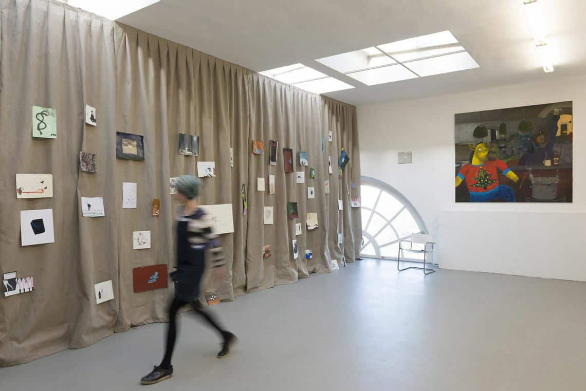 Foto: Ein Raum mit weißen Wänden, Bogen- und Deckenfenstern. Durch den Raum läuft ein deckenhoher Vorhang in einem beigeton, auf dem viele bemalte Papiere auf unterschiedlicher Höhe angebracht sind. An der Wand hängt eine großformatige Leinwand. Durch den Raum bewegt sich ein Mensch.