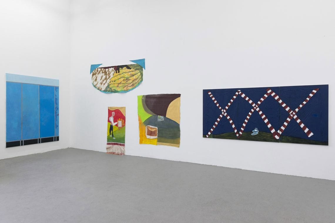 Foto: Ein Raum mit weißen Wänden und grauem Boden. An den Wänden hängen mehrere flache Objekte in Groß- und Mittelfromaten neben- und übereinander.