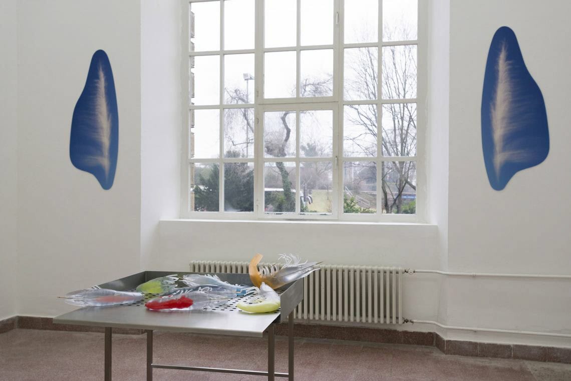 Foto: Ein Raum mit weißen Wänden, rotem Steinboden und einem großen Fenster. Links und rechts neben dem Fenster hängen Objekte, die einander entsprechen, jedoch gespiegelt sind. Im Raum steht ein weiteres Objekt, das aus unterschiedlichen Materialien besteht.