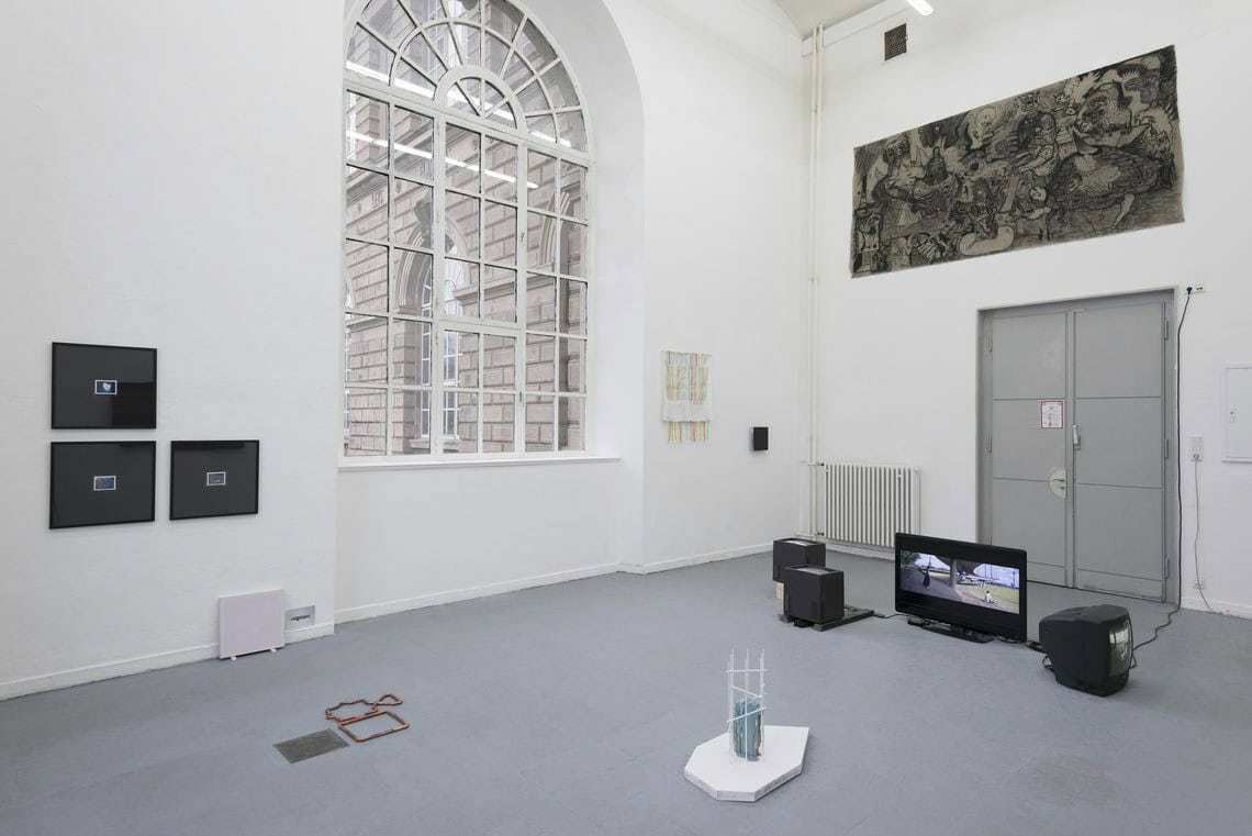 Foto: Ein Raum mit weißen Wänden, grauem Boden, großen Fenster und einer zweiflügligen Tür. An den Wänden hängen eine Vielzahl von Objekten auf unterschiedlicher Höhe. Auf dem Boden liegen und stehen Skulpturen und mehrere Bildschirme.